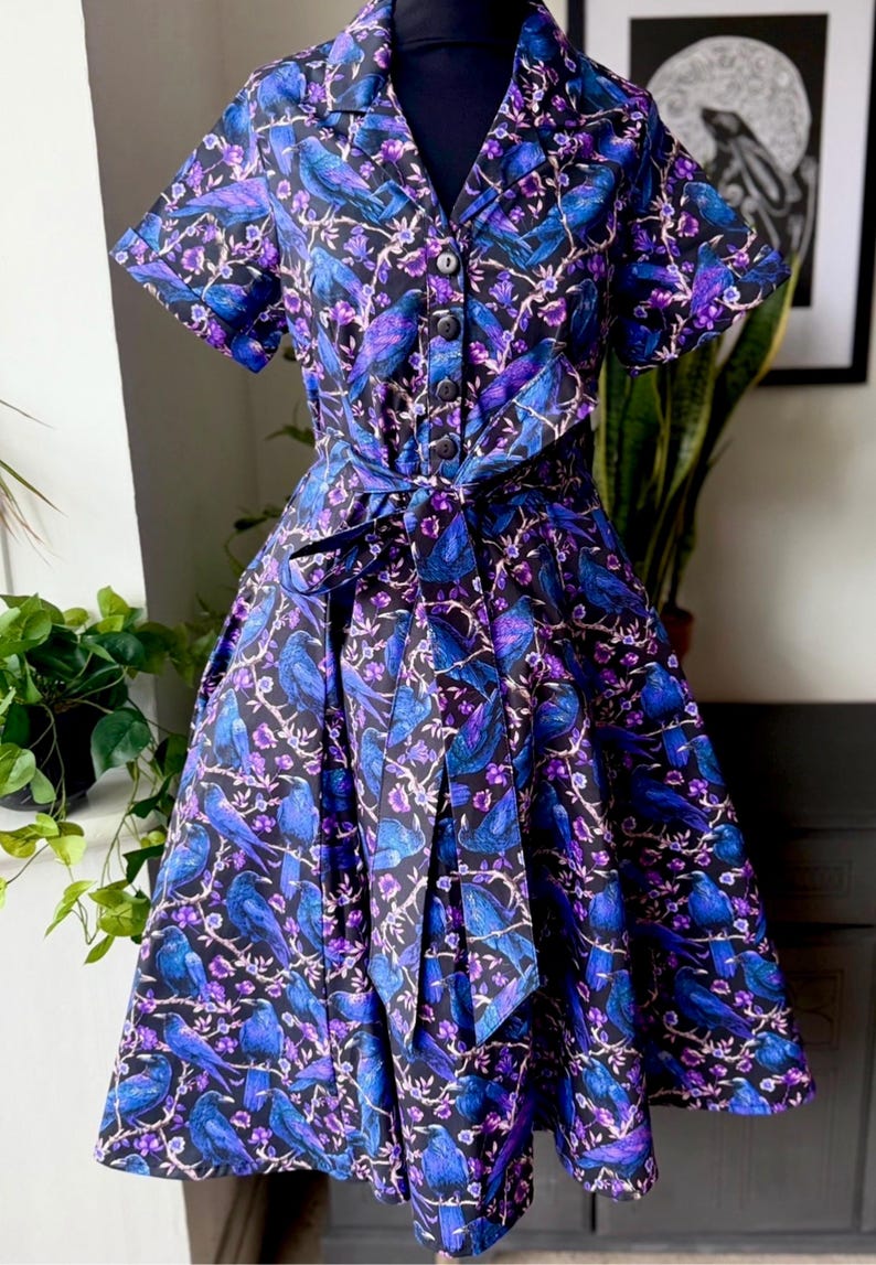 May include: A short-sleeved dress with a black background, featuring a pattern of blue birds and purple flowers. The dress has a collared neckline, a button-down front, and a matching tie belt. The skirt is pleated.
