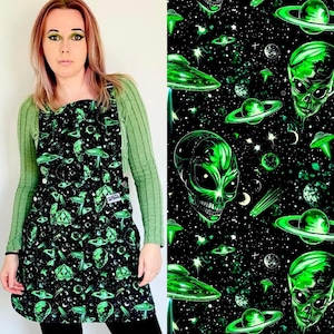 Può includere: Grembiule nero con un motivo a tema alieno e spazio verde neon. Il grembiule presenta un motivo ripetuto di teschi alieni, UFO, pianeti e stelle. Il grembiule è indossato sopra una camicia verde a maniche lunghe.