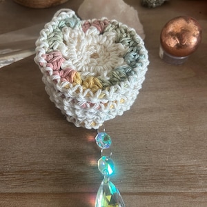 Può includere: Un sottobicchiere all'uncinetto bianco fatto a mano con accenti pastello di rosa, giallo e verde. Un prisma di cristallo decorativo è appeso sotto, riflettendo la luce. Un oggetto di arredamento per la casa unico.