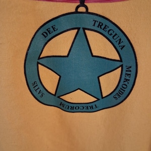 May include: A mustard yellow t-shirt featuring a dark teal circular pendant graphic with a central star. The circular border contains the black text DEE TREGUNA MEKOIDES SATIS TRECORUM. The shirt has a dark red collar trim at the top neckline.