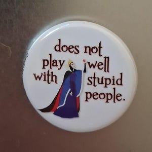 May include: A round white button featuring a graphic of a queen in a purple and black gown with a red cape. The text printed in dark red reads: does not play well with stupid people. The design is centered on the circular surface of the button.