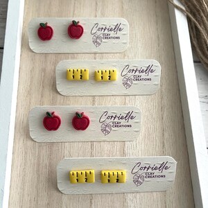 Op de afbeelding: Vier paar polymer clay oorbellen op witte kaartjes. Twee paar zijn rode appel oorbellen en twee paar zijn gele liniaal oorbellen. Elk kaartje heeft de tekst "Corrielle Clay Creations".