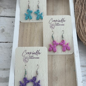May include: Three pairs of balloon dog earrings on display cards. The earrings are made of polymer clay and come in teal, pink, and purple.