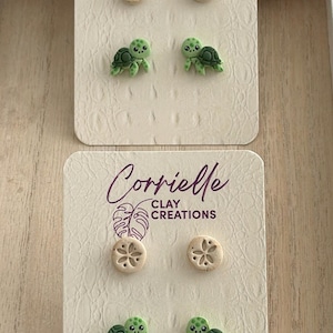 May include: Two sets of stud earrings on branded Corrielle Clay Creations display cards. Each set includes a pair of beige circular sand dollar studs and a pair of light green turtle studs with dark green shell details and black eyes.