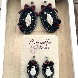 Op de afbeelding: Handgemaakte Halloween-oorbellen met een spookontwerp. De oorbellen zijn zwart met een witte geest, rode rozen en een strik. De oorbellen staan op een witte kaart met de tekst "Corrielle Clay Creations".