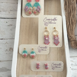 May include: A set of five pairs of polymer clay earrings featuring summer-themed designs. The earrings are displayed on white cards with the brand name "Corrielle Clay Creations". The designs include a pair of flip-flop earrings, a pair of swimsuit earrings, and three pairs of earrings with a beach scene.