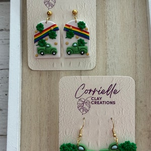 May include: Two pairs of green truck earrings with shamrocks and rainbows. The earrings are on white cards with the text "Corrielle Clay Creations".