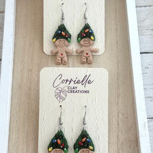 May include: A pair of dangle earrings featuring a troll doll wearing a green Christmas tree hat with colorful lights. The earrings are on a white card with the text "Corrielle Clay Creations".
