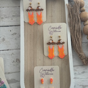May include: Three sets of polymer clay earrings featuring a swimsuit, a hat, and flip-flops. The earrings are on white cards with the text "Corrielle Clay Creations".