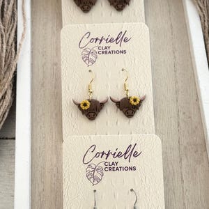 May include: Three pairs of dangle earrings featuring brown highland cow heads with floral accents. The top pair has pink flowers, the middle pair has yellow flowers, and the bottom pair has pink flowers. The earrings are displayed on white cards with the text "Corrielle Clay Creations".