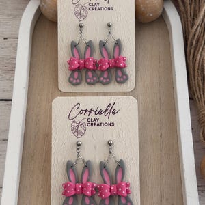 Puede incluir: Pendientes grises con forma de conejito con lazos rosas y lunares blancos, colgados de cadenas plateadas. Se exhiben en una tarjeta crema con el texto "Corrielle Clay Creations". Un accesorio encantador y juguetón.