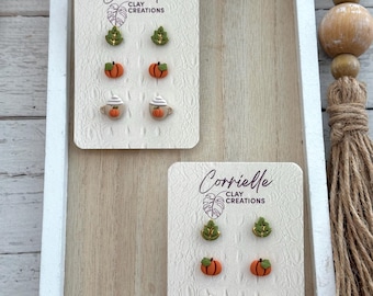 May include: A set of six fall-themed stud earrings featuring a pumpkin, a coffee cup with whipped cream, and a green leaf. The earrings are displayed on a white card with the text "Corrielle Clay Creations".