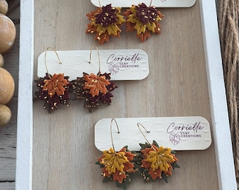 Pode incluir: Cinco pares de brincos de argola com pingentes de folhas de bordo de argila em camadas em cores de outono como bordô, amarelo mostarda, laranja e verde escuro, acentuados com flocos de folha de ouro. Cada par está preso a um cartão branco com a marca.