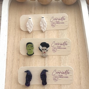 Op de afbeelding: Drie paar Halloween-thema polymer clay oorbellen getoond op een houten oppervlak. De eerste set heeft witte spook oorbellen, de tweede set heeft groene Frankenstein en zwart-witte Bride of Frankenstein oorbellen, en de derde set heeft zwarte kraai oorbellen. Elke set is gelabeld met de tekst "Corrielle Clay Creations".