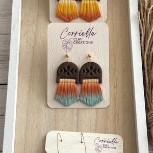 May include: Earrings with a dark brown arch design and a fringe of yellow, orange, and blue. The earrings are displayed on a white card with the text "Corrielle Clay Creations". The image also shows pumpkin-shaped earrings.
