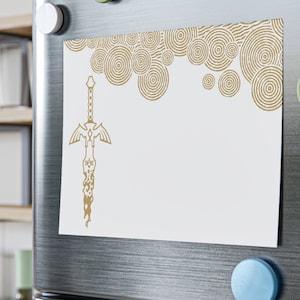 May include: A white refrigerator magnet with a gold sword design and a background of gold circles. The magnet is on a stainless steel refrigerator.