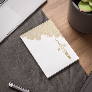 May include: A white notepad with a gold design of a sword and a pattern of circles. The notepad is on a gray surface.