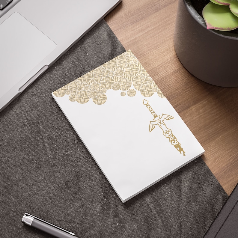 May include: A white notepad with a gold design of a sword and a pattern of circles. The notepad is on a gray surface.