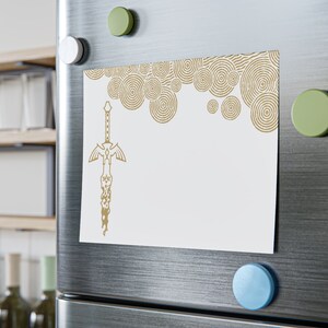 May include: A white refrigerator with a gold and white decorative magnet featuring a stylized sword design. The magnet has a pattern of concentric circles in the background.