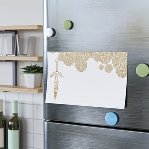 May include: A white refrigerator with a gold sword and circle design magnet on the door. The magnet has a white background with a gold sword and a pattern of overlapping circles.