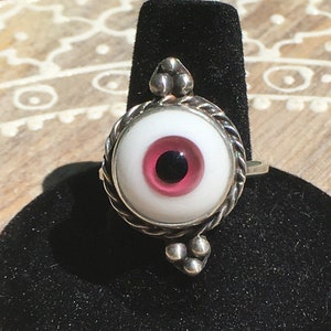 May include: A silver ring with a white and pink eye design. The eye has a black pupil and a pink iris.