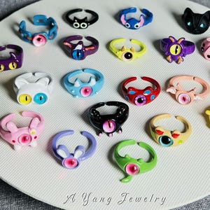 May include: A collection of colorful, adjustable rings with cartoon monster and animal designs. The rings feature various colors, including pink, blue, yellow, and black, with detailed eye and face features. The rings are displayed on a white surface.