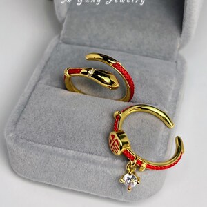 Peut inclure: Bagues dorées avec des détails rouges et une pierre précieuse transparente. L'une des bagues est un motif de serpent, l'autre a un charme avec un caractère chinois et une pierre précieuse pendante. Les bagues sont présentées dans un écrin en velours gris. Le texte sur la boîte indique "A Yang Jewelry".