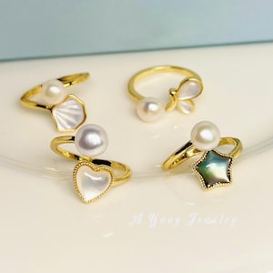 May include: Four gold-toned rings with pearl accents and decorative elements. One ring features a seashell design, another a butterfly, a third a heart, and the last a star-shaped iridescent stone. The rings are displayed on a white surface.