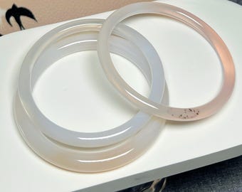 May include: Three translucent, light-colored bangles are displayed on a white surface. The bangles vary in shades of white and pink. The text "A Yang Jewelry" is visible at the bottom of the image. The bangles are a type of jewelry.