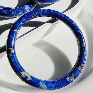 May include: A Yang Jewelry. Close-up of three blue lapis lazuli bangles. The bangles are a deep blue color with white and gold veining. The text "A Yang Jewelry" is at the bottom of the image.