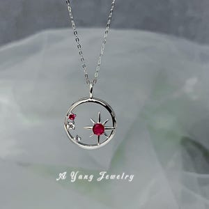 May include: A silver necklace featuring a pendant with a celestial design. The pendant has a circular frame with a starburst and two red gemstones. The necklace is delicate and elegant, perfect for everyday wear. The text "A Yang Jewelry" is at the bottom.