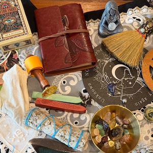 May include: A collection of items including a Tattoo Tarot deck, a red leather journal, a small broom, a wooden pentacle, a brass bowl filled with assorted crystals, a sage bundle, wax sealing sticks, a stamp, a feather, and a small figurine.