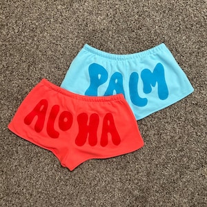 Puede incluir: Dos pares de pantalones cortos. Un par es de color rojo brillante con la palabra "Aloha" en letras grandes a juego. El otro par es de color azul claro con la palabra "Palm" en letras grandes de color azul oscuro. Ambos pares tienen cintura elástica.