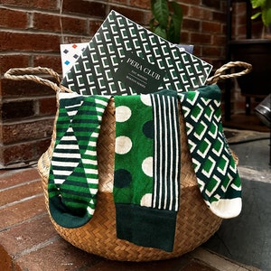 May include: A wicker basket filled with three pairs of green and white patterned socks. The socks are folded and arranged in the basket. The basket is sitting on a brick surface. The socks are labeled "PERA CLUB EST. BOSTON BOSTON SERIES".