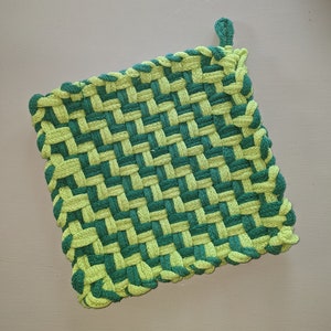 Puede incluir: Un salvamanteles cuadrado tejido en tonos verdes y verde lima. El salvamanteles está hecho de hilo grueso, con una esquina en bucle para colgarlo. El patrón de tejido es un diseño apretado, similar a una cesta.