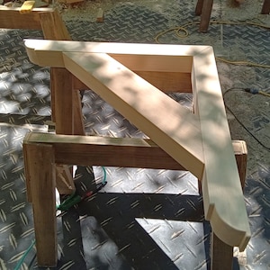 May include: A wooden corner brace with a rounded edge, resting on two sawhorses. The brace is made of light-colored wood and has a smooth finish.