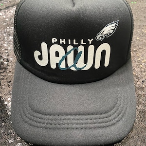 Op de afbeelding: Zwarte truckercap met een wit "PHILLY JAWN" logo en een wit Philadelphia Eagles logo. De pet heeft een zwart mesh achterpaneel.