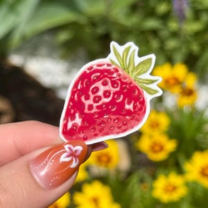 May include: A red strawberry sticker with green leaves. The sticker is held up against a blurred background of yellow flowers and green foliage. The strawberry has a detailed design.