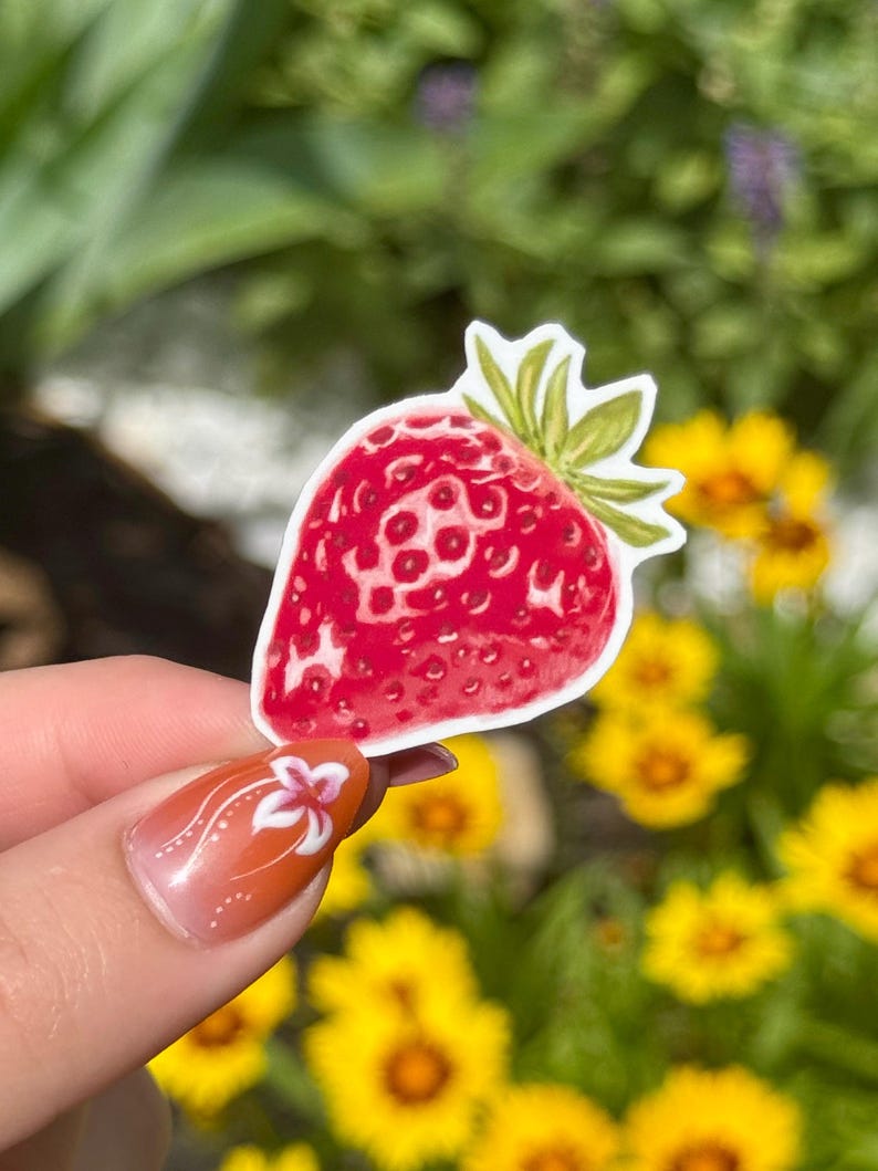 May include: A red strawberry sticker with green leaves. The sticker is held up against a blurred background of yellow flowers and green foliage. The strawberry has a detailed design.