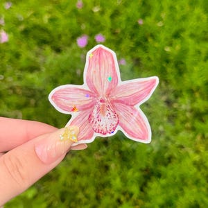 May include: A pink orchid sticker with a white border, held against a blurred green background. The sticker has a detailed watercolor design with a speckled center and iridescent glitter accents. The flower's petals are a soft pink.