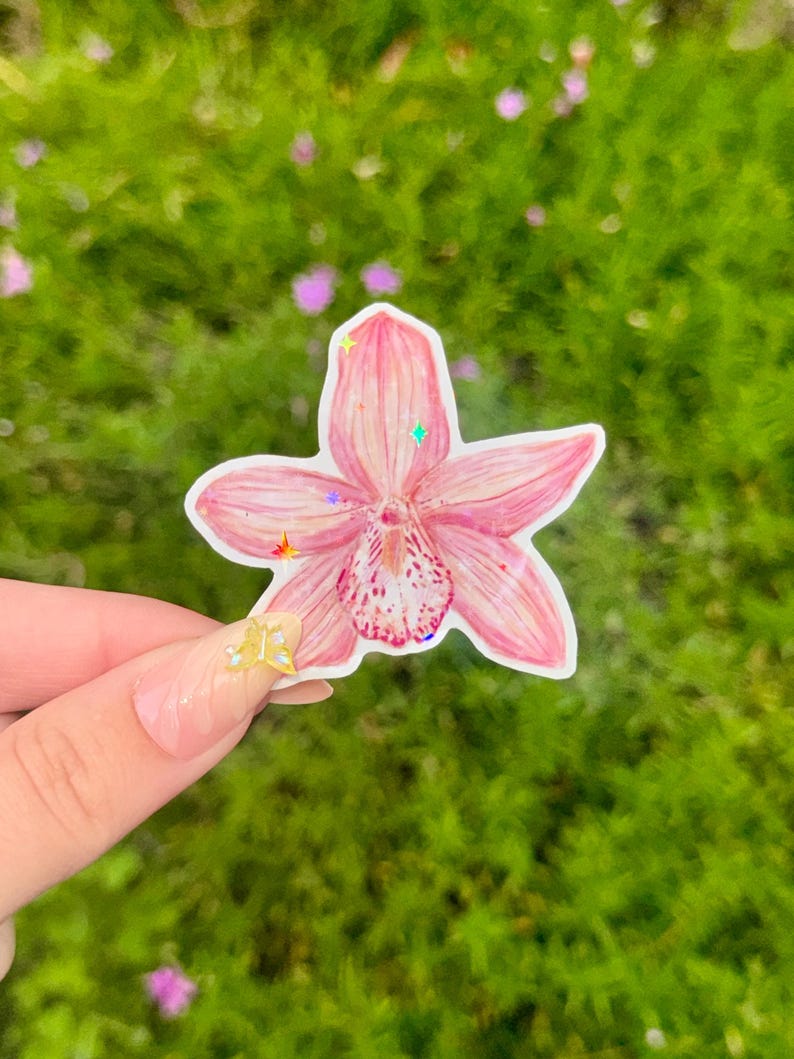 May include: A pink orchid sticker with a white border, held against a blurred green background. The sticker has a detailed watercolor design with a speckled center and iridescent glitter accents. The flower's petals are a soft pink.