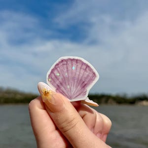 May include: A purple seashell sticker with iridescent glitter stars. The sticker is being held by a hand with a flower nail design.