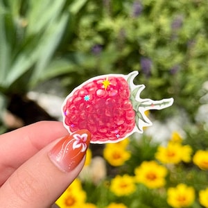 May include: A red raspberry sticker with a green stem and glitter accents. The sticker is held in front of a blurred background of yellow flowers and green foliage.