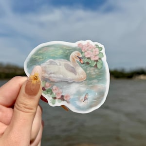 May include: A white swan sticker with pink flowers and green leaves on a blue and green watercolor background. The sticker is on a hand holding it up against a blue sky and water.