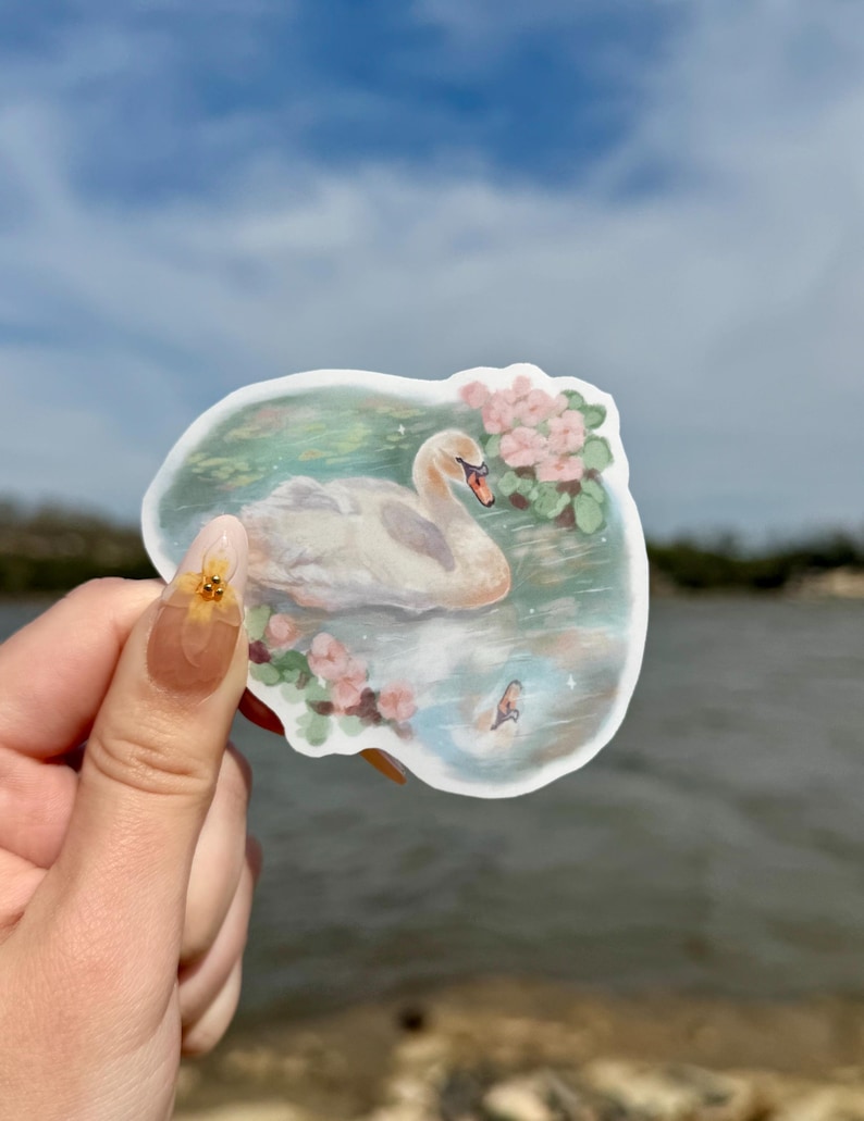 May include: A white swan sticker with pink flowers and green leaves on a blue and green watercolor background. The sticker is on a hand holding it up against a blue sky and water.