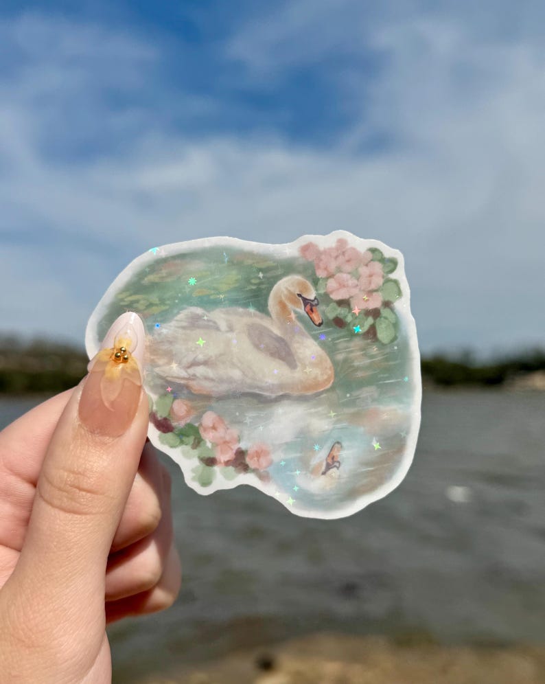 May include: A white swan sticker with a glittery finish, surrounded by pink flowers and green leaves. The sticker is on a white background and has a slightly blurred background of a body of water.