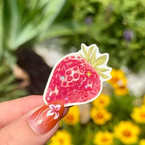May include: A red strawberry sticker with green leaves and glitter accents. The sticker is held up against a blurred background of green foliage and yellow flowers.