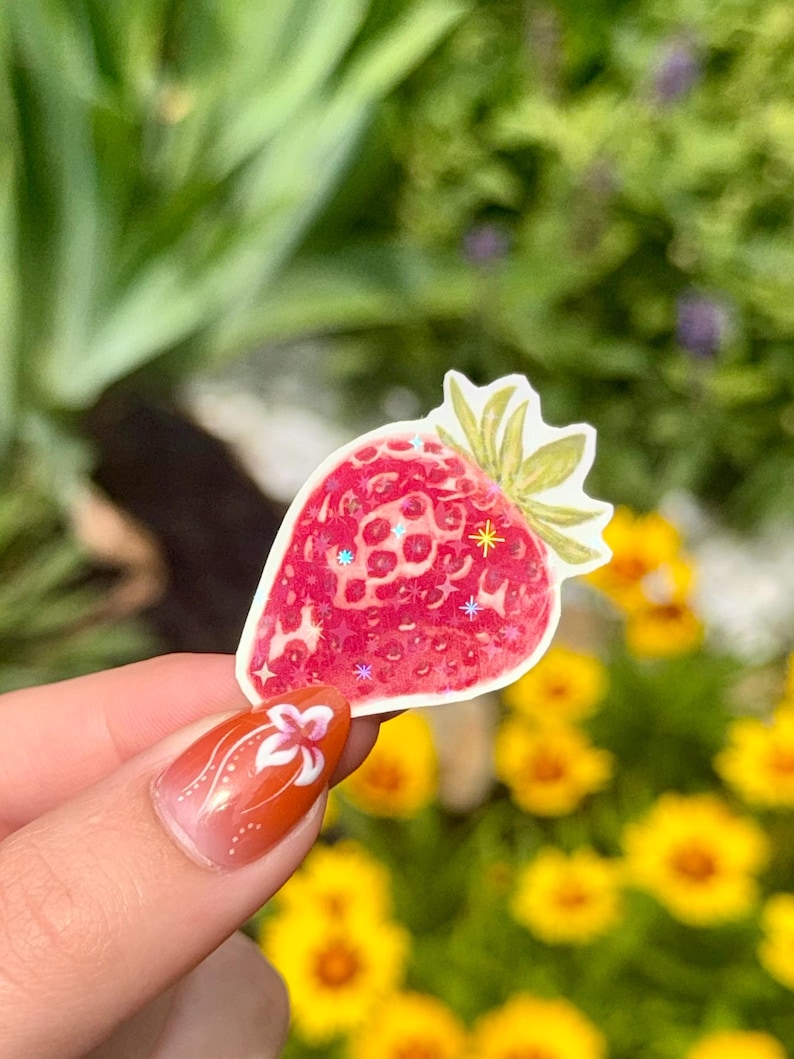 May include: A red strawberry sticker with green leaves and glitter accents. The sticker is held up against a blurred background of green foliage and yellow flowers.