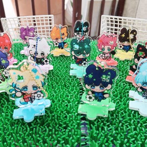 Puede incluir: Una colección de llaveros acrílicos coloridos con personajes de estilo anime con uniformes de fútbol. Cada llavero tiene un color de pelo y diseño únicos, con un balón de fútbol. Los llaveros están sobre una superficie verde con una portería de fútbol en miniatura en el fondo.