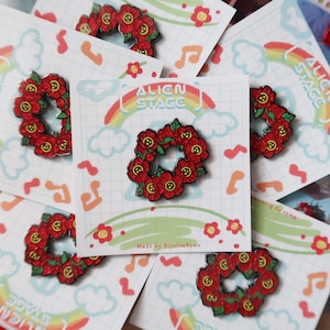 May include: A collection of enamel pins featuring a red floral wreath design. The pins are on cards with the text "Alien Stage" and a rainbow graphic. The background includes musical notes and flowers.